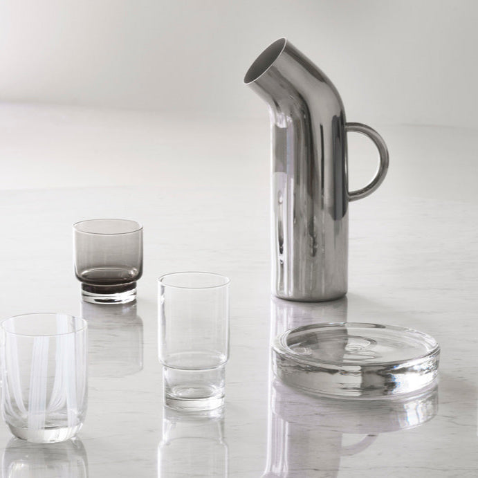 Clear glass pitcher and tumblers on a reflective surface with a light gray background