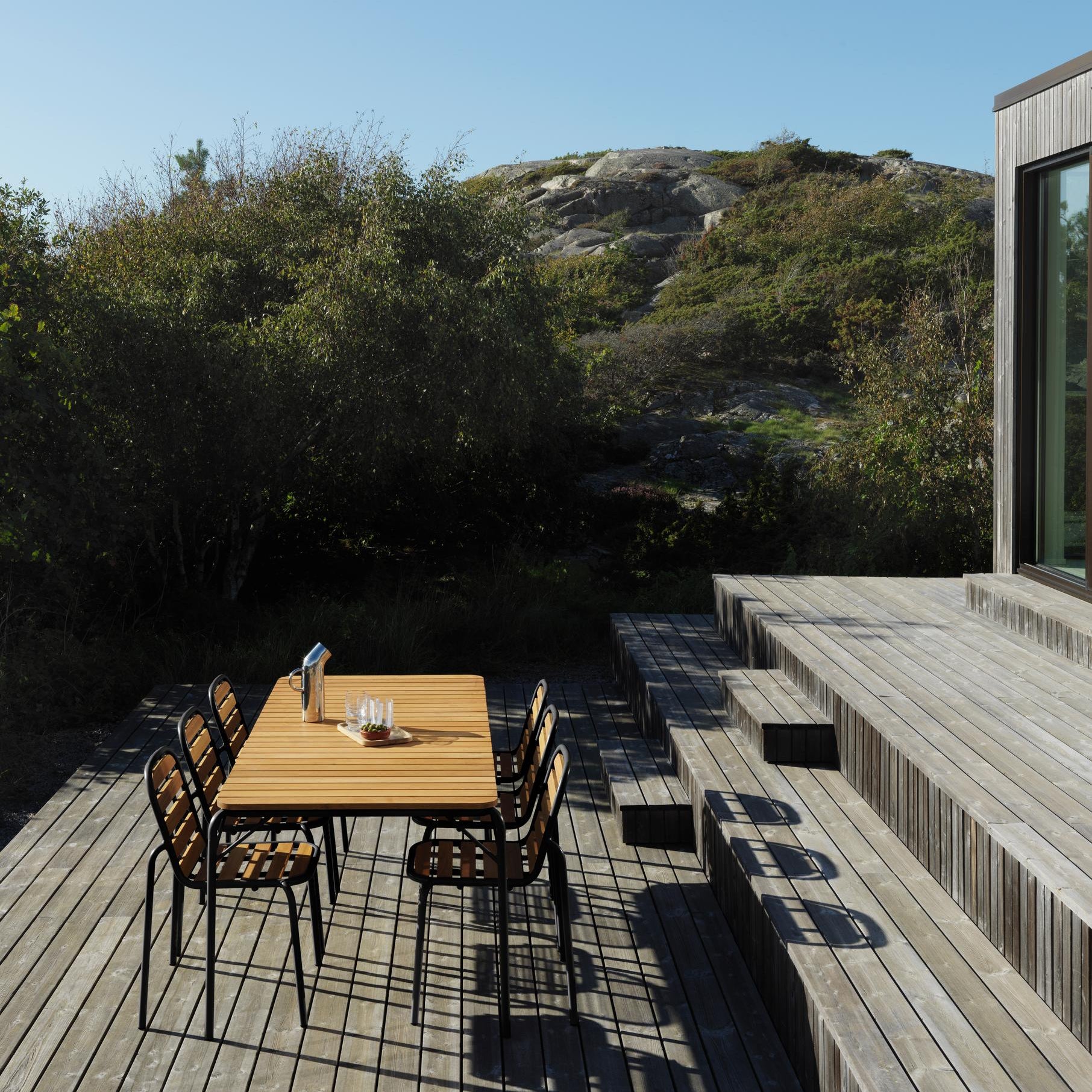 Outdoor wooden dining area with chairs on a deck, surrounded by nature.