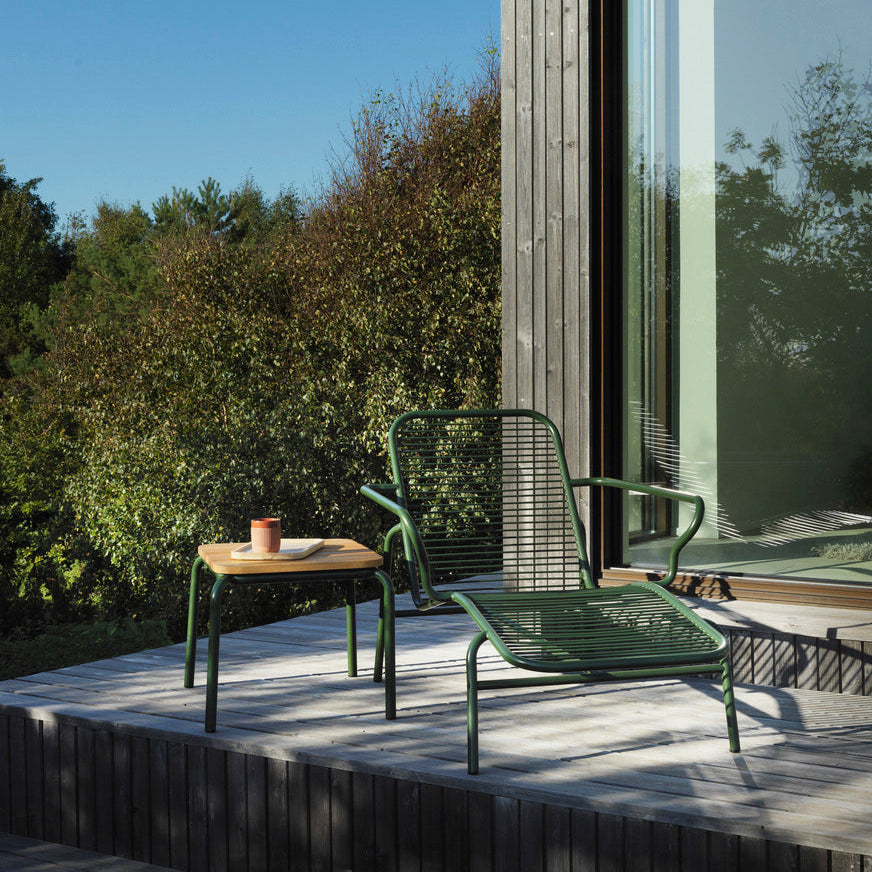 Modern outdoor setting with a green chair and table on a wooden deck, surrounded by trees and a clear blue sky.
