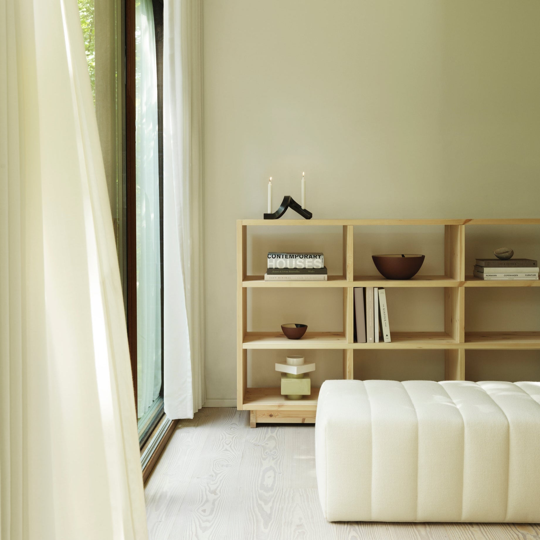 Modern interior with a wooden bookshelf and white ottoman near a large window.