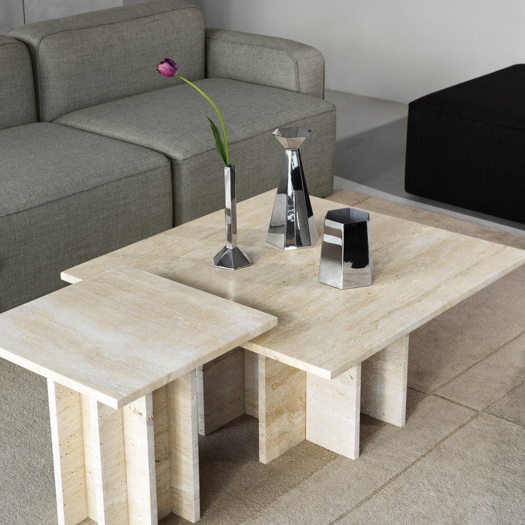 A modern multi-piece nesting coffee table set made of light beige travertine stone, styled in a living room next to a gray fabric sofa with HEXA stainless steel vases.