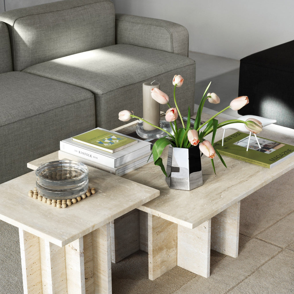 A modern multi-piece nesting coffee table set made of light beige travertine stone, styled in a living room next to a grey fabric sofa with a set of decorative stainless steel vases.