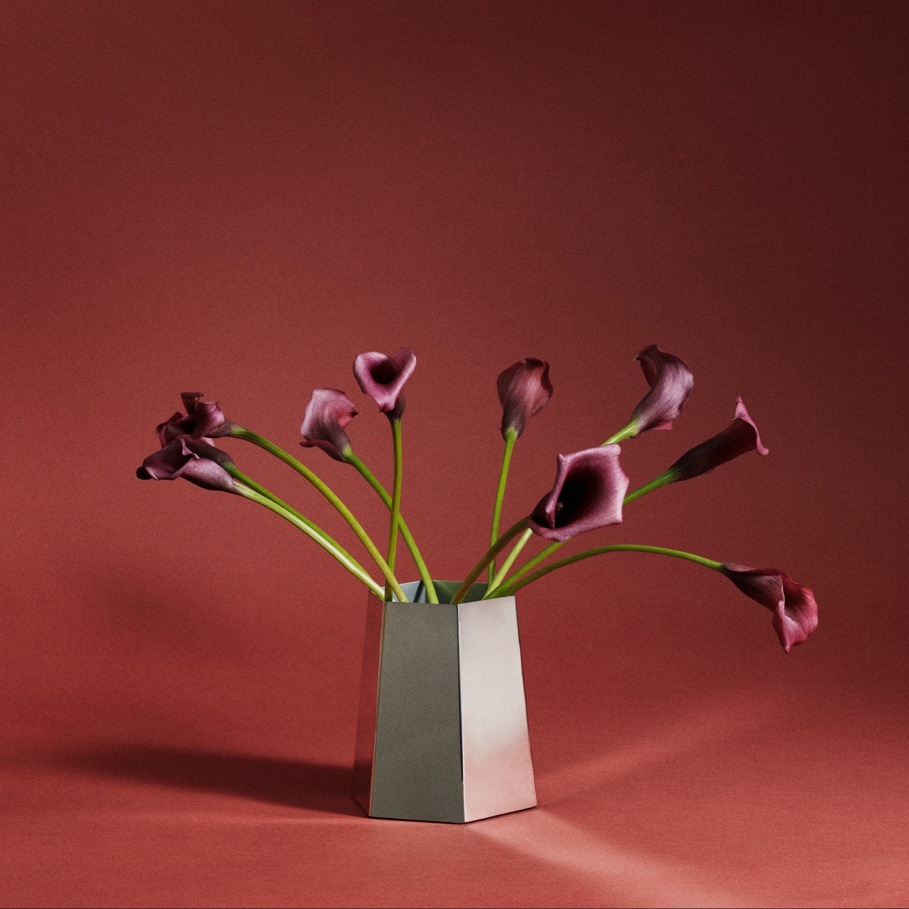 A modern mirrored stainless steel geometric vase holding a bouquet of dark purple calla lilies, set against a solid dark reddish-brown studio background.