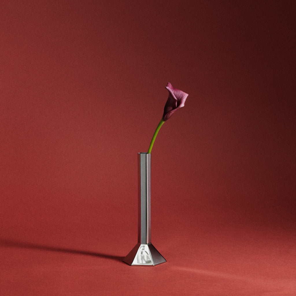 A single pink calla lily flower in a tall, single stem stainless steel vase with a geometric base, set against a solid dark red studio background.