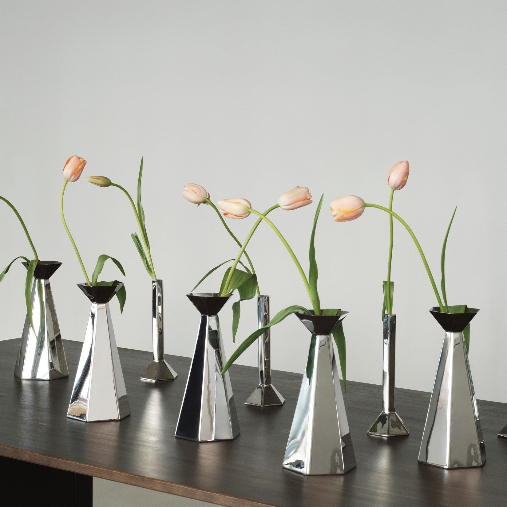 A set of modern metallic silver tulip-shaped vases arranged on a dark wooden table against a light gray wall, each holding a single pale pink tulip.