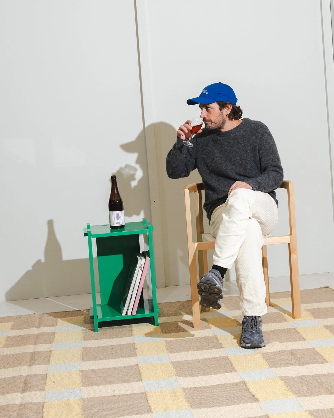 Person sitting on a chair with a bottle and books on a small table, wearing a blue cap and dark sweater.