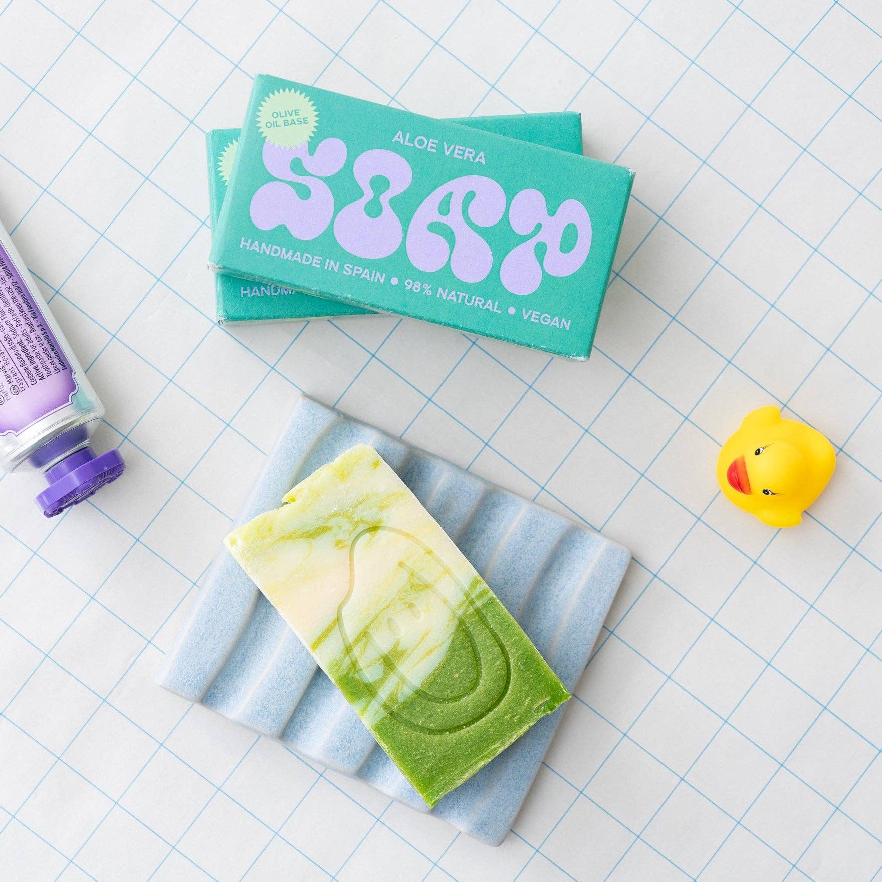 Bar of soap with green swirls on a striped towel, next to a box labeled 'Aloe Vera' on a grid background.