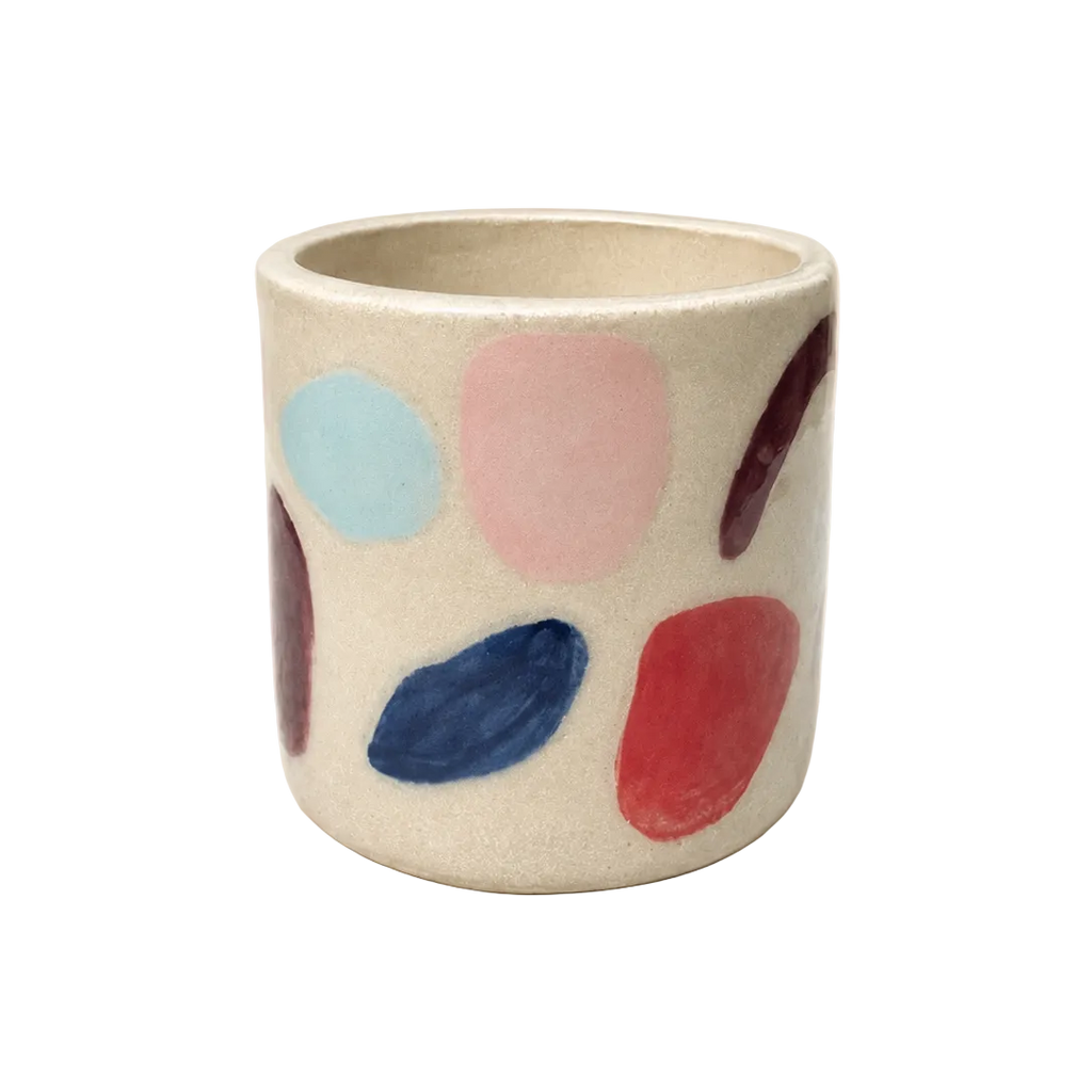 A small cylindrical ceramic cup with a textured off-white glaze, decorated with hand-painted abstract polka dots in light blue, pink, navy, red, and dark purple, set against a solid cream background.