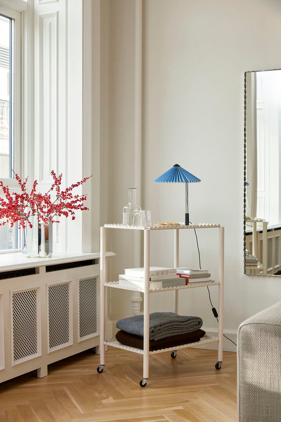 White rolling cart with books and a lamp in a room with large windows and a radiator.