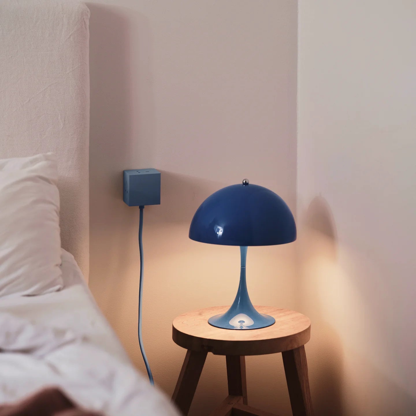 Blue lamp on a wooden stool next to a bed with a white pillow.