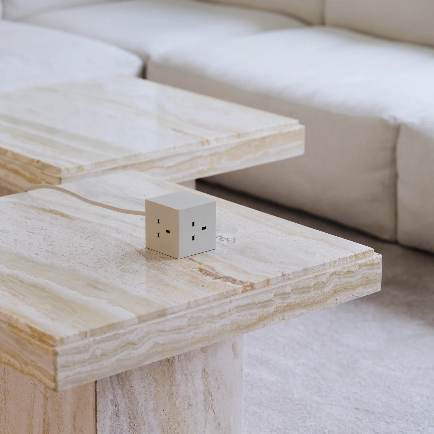A small white cube-shaped electrical outlet socket with UK-style three-pin plugs sits on a light beige marble coffee table in a modern living room setting with a matching sofa in the background.