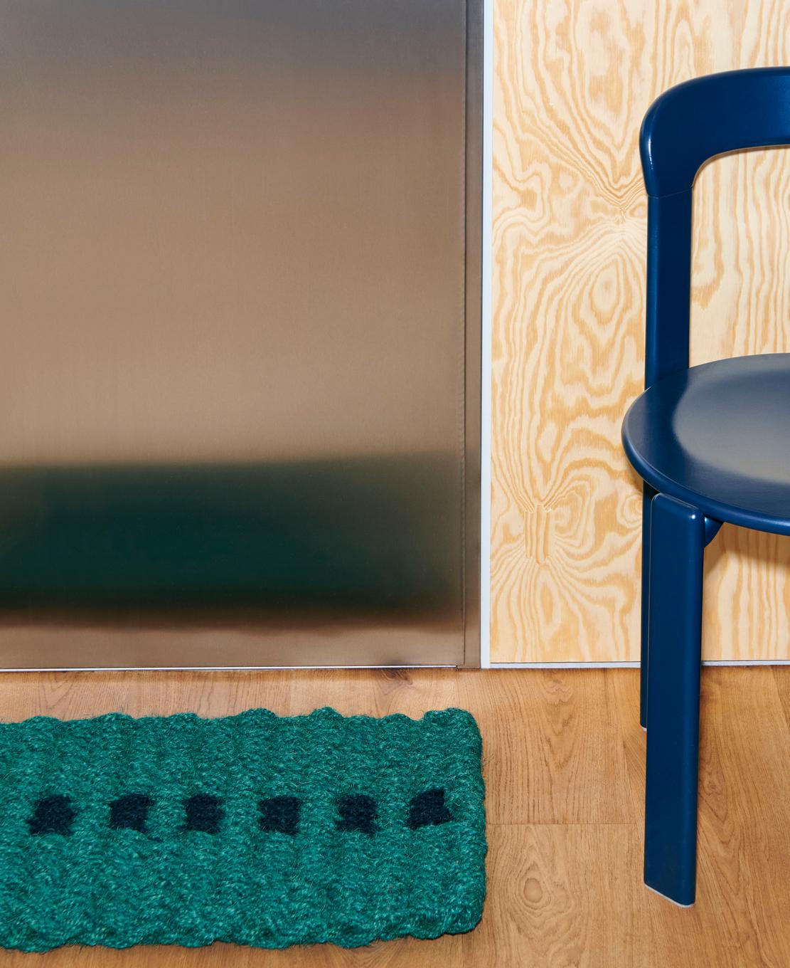 Teal doormat with black dots on a wooden floor next to a blue chair.