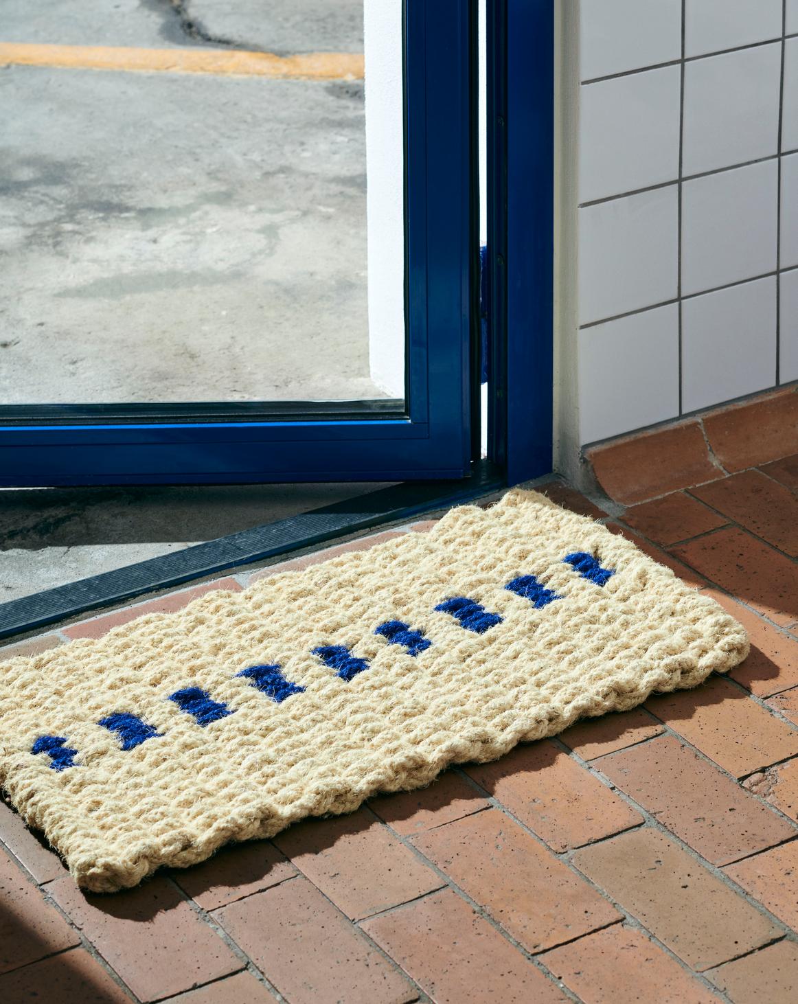 Doormat with blue text on a tiled floor outside a glass door.