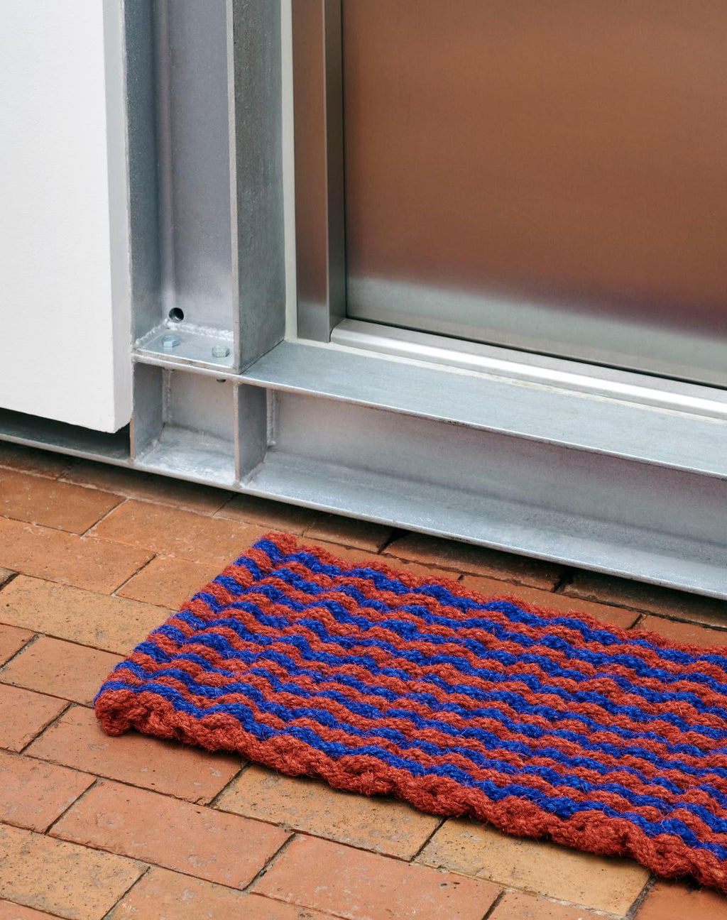 Red and blue doormat on a brick floor in front of a glass door.