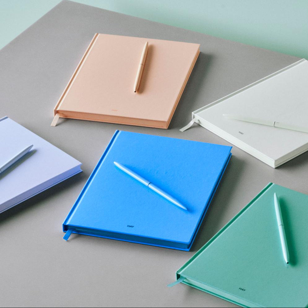 Six hardcover notebooks in pastel and neutral colors arranged on a gray surface against a mint green background, each with a matching pen resting on top.