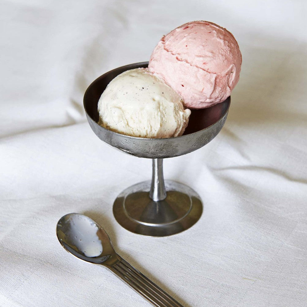 Two scoops of ice cream in a metal bowl on a white surface with a spoon.