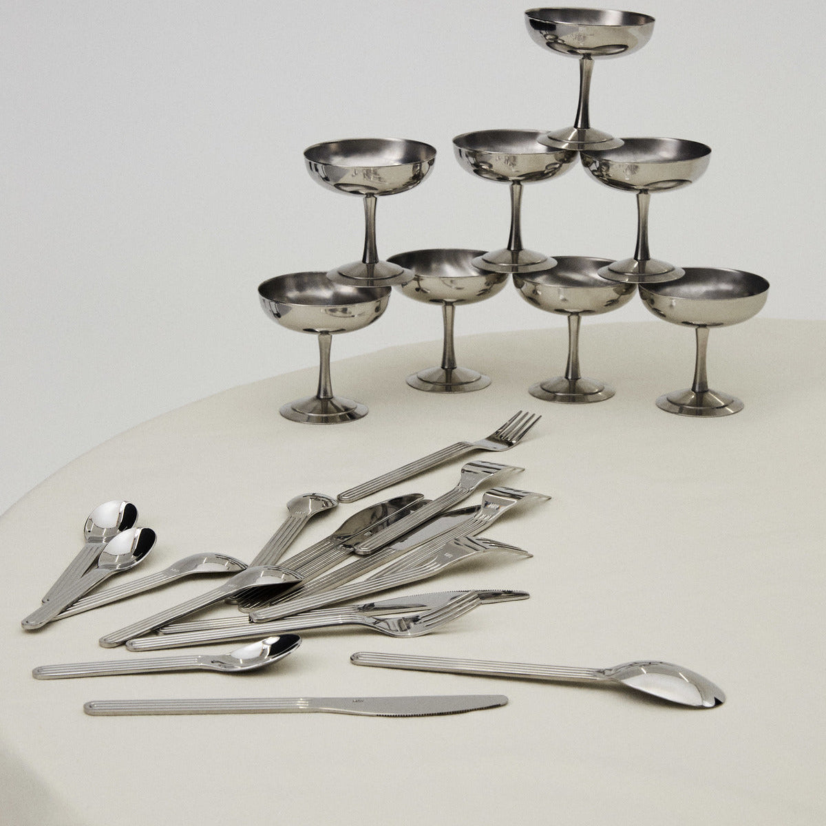 Set of silver cutlery including spoons, forks, and knives on a white surface with small metallic bowls arranged in a pyramid formation.