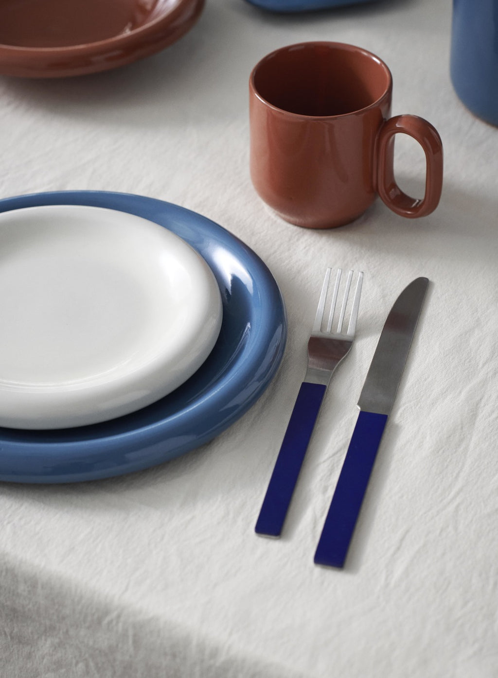 Ceramic tableware including a blue plate, white plate, red mug, fork, and knife on a light gray surface.