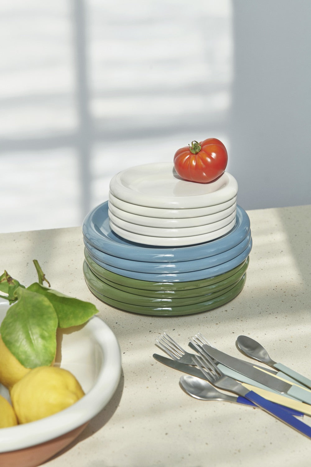 Stacked ceramic plates in blue, green, and white on a kitchen counter with utensils and fruit.