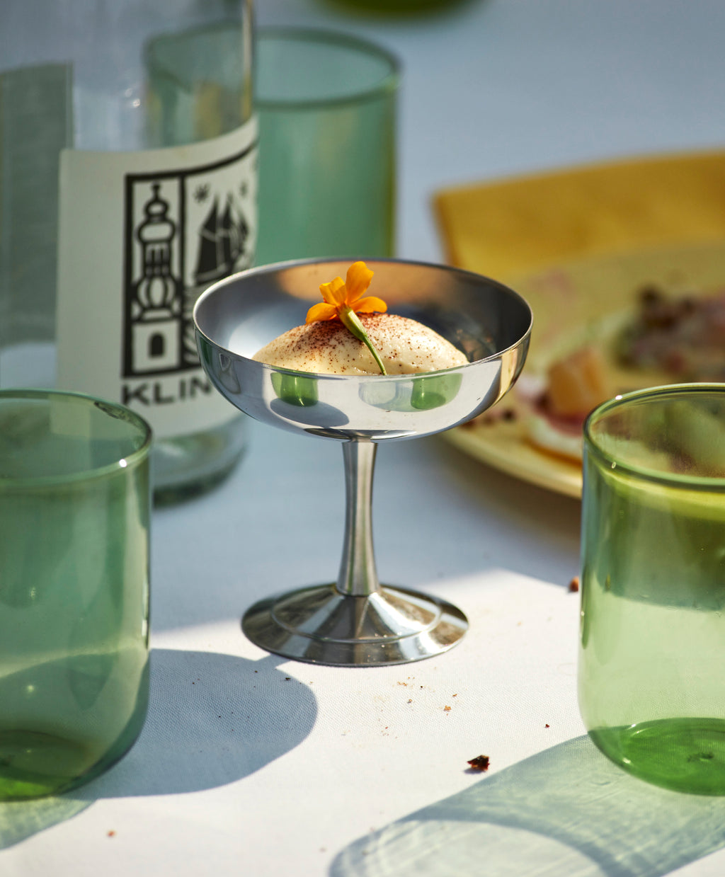 Metallic cocktail glass with a small dish and garnish on a table with green glasses and a blurred background.