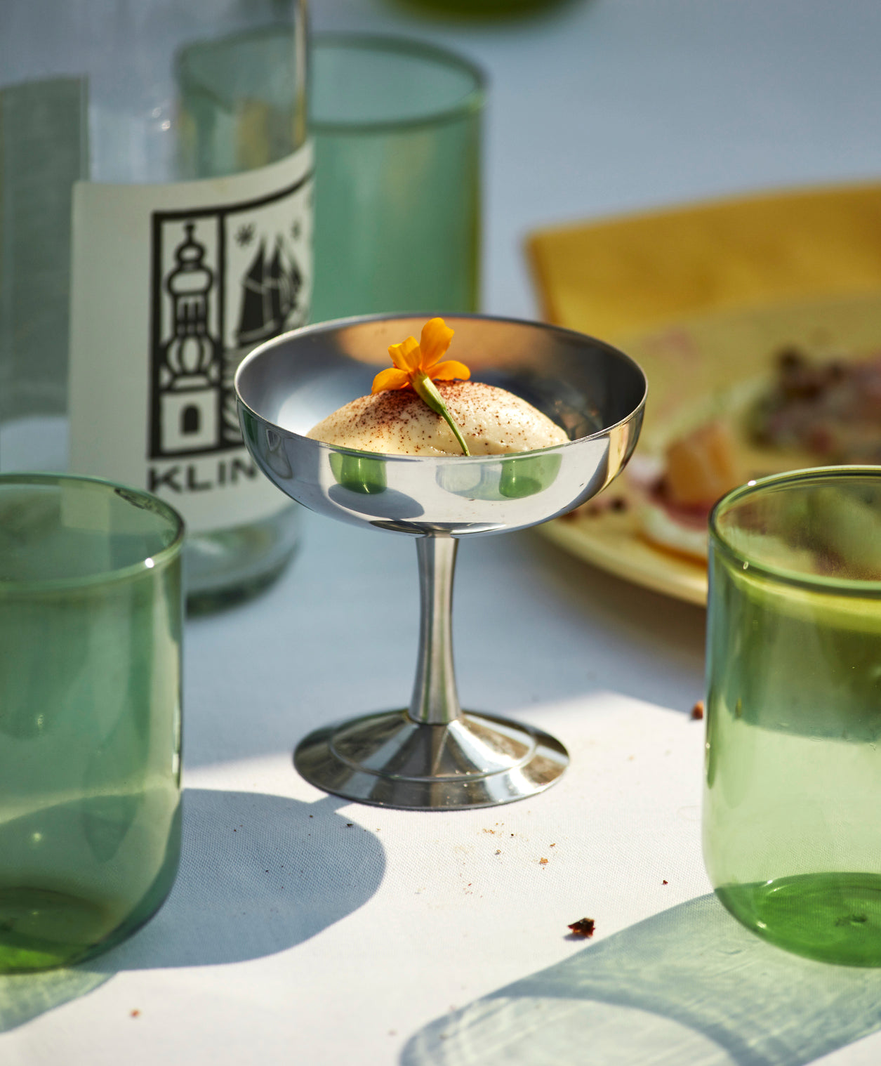 Metallic cocktail glass with a small dish and garnish on a table with green glasses and a blurred background.