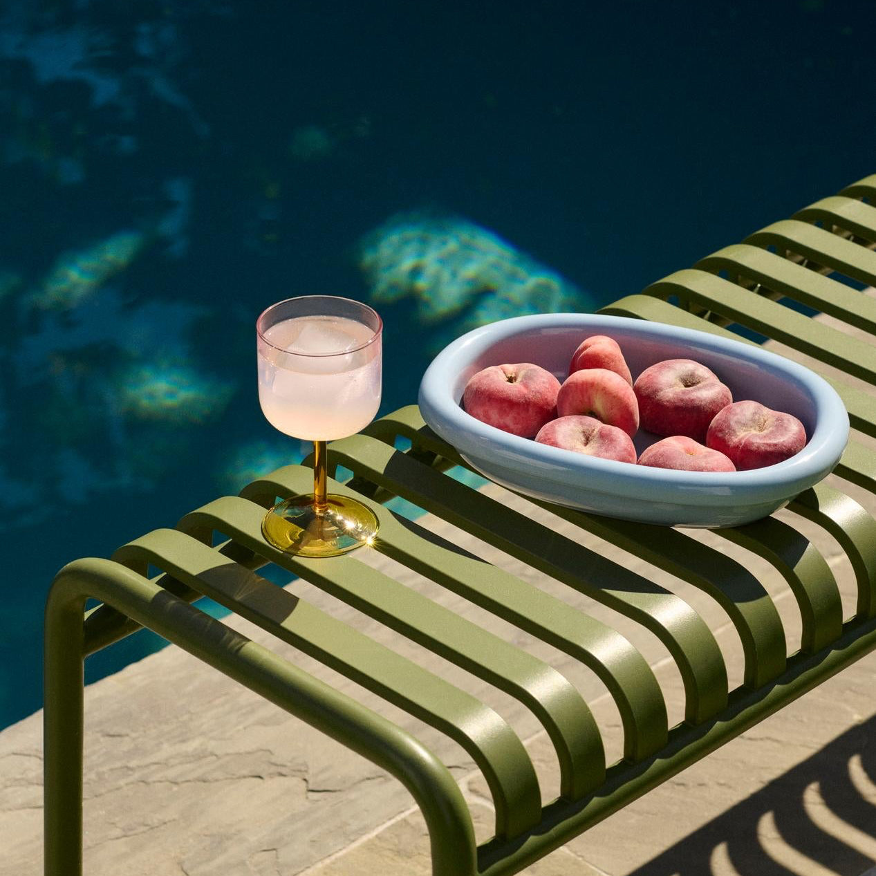 Green metal bench with a bowl of apples and a glass of pink drink by a pool.