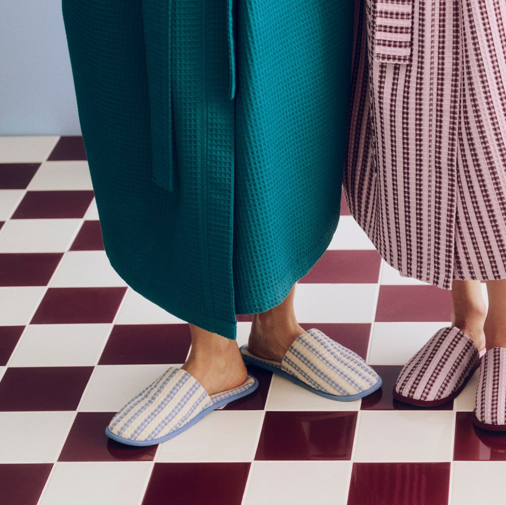 Three people standing on a red and white checkered floor, wearing matching striped bathrobes and coordinated slip-on slippers in teal, pink, and burgundy tones.