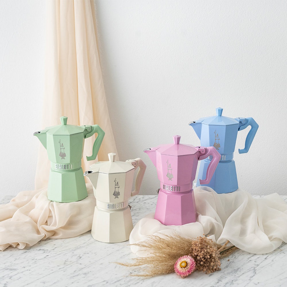 Four colorful coffee percolators on a light surface with a neutral curtain background