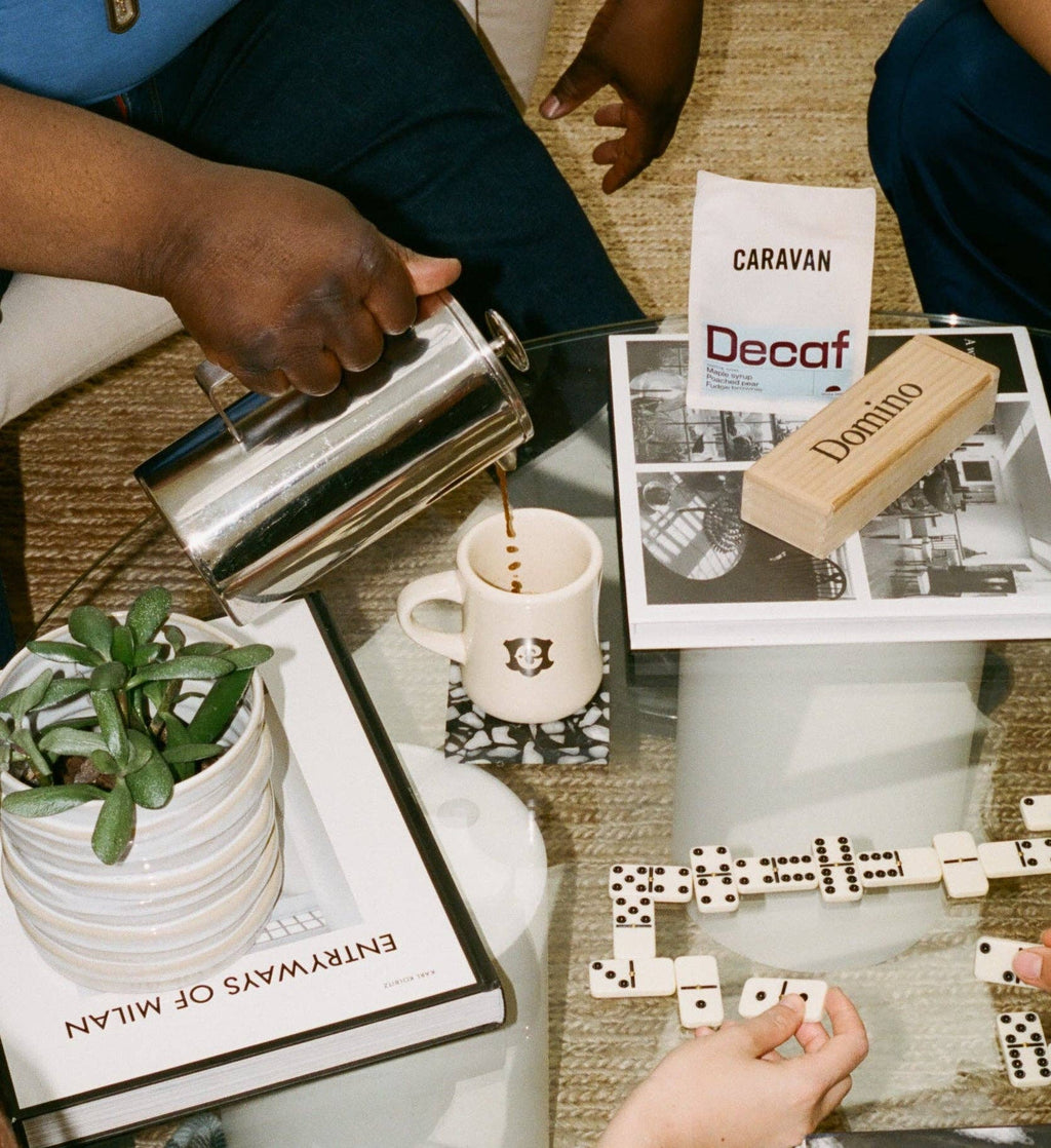 Person pouring coffee from a French press on a table with Domino and Caravan coffee bags, a plant, and dominoes.